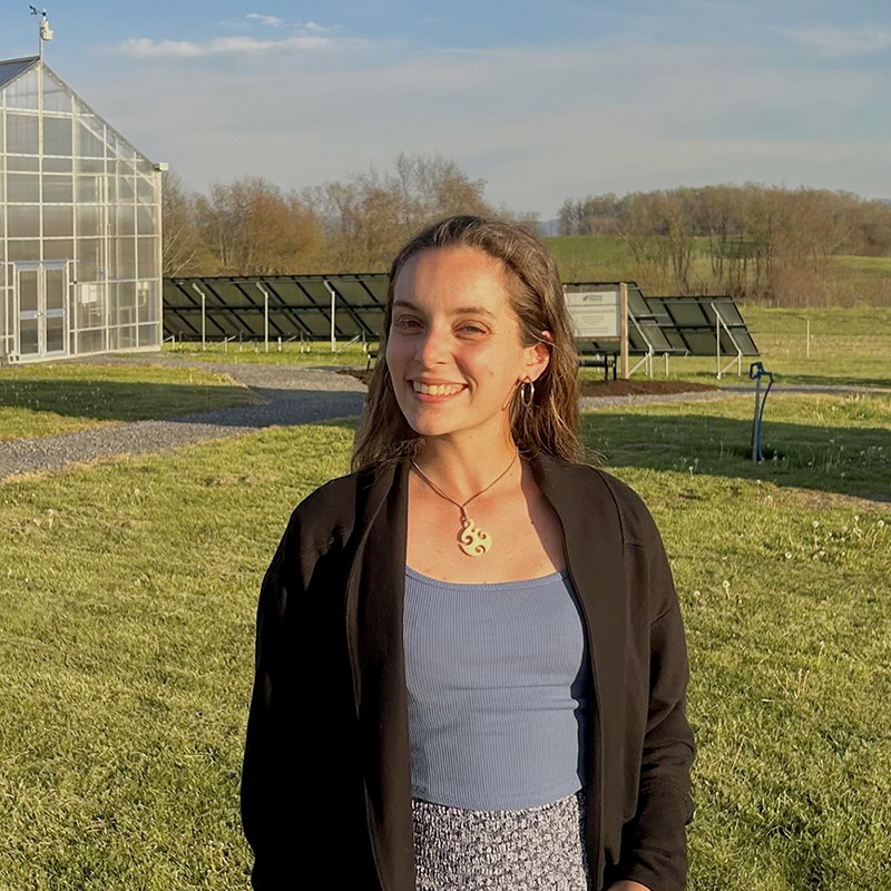 Young woman smiling with a greenhouse building in the background.