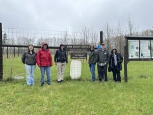Several people stand in the raing around a sign that reads Tom Rush Forest. In the background is an open field with chestnut trees planted in it.