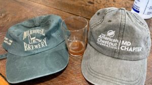 A Milkhouse Brewery ball cap next to a pint of lager next to a MD Chapter of TACF ball cap.
