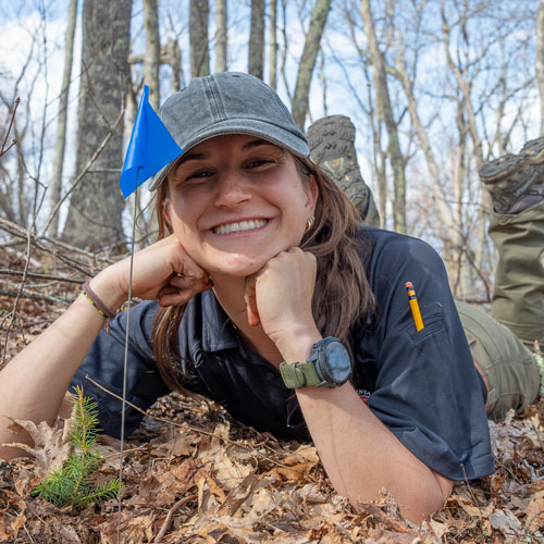 Zoe Bergman in ball cap and outdoor wear posing next to a tiny spruce planting.