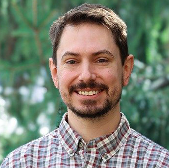 Steve Morrone headshot. Man with dark hair, mustache and beard. He wears a plaid shirt and has greenery in the background.